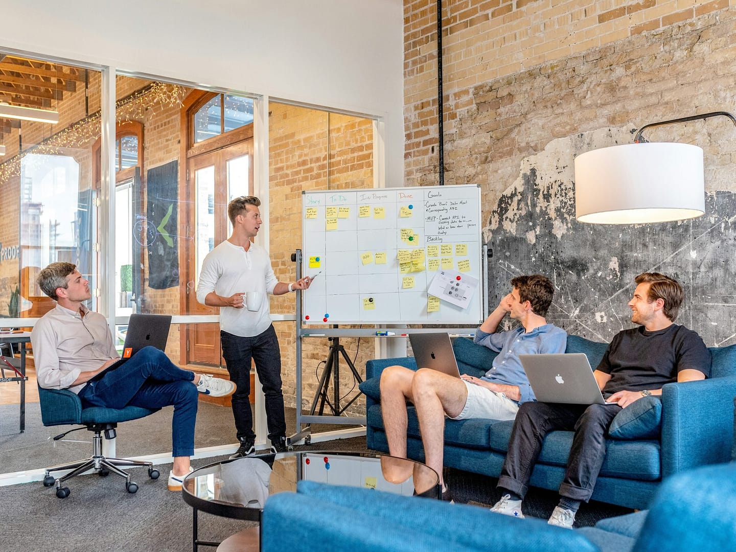 A group of four colleagues in a modern office engaged in a strategy session, with one person presenting ideas on a whiteboard and others seated with laptops.