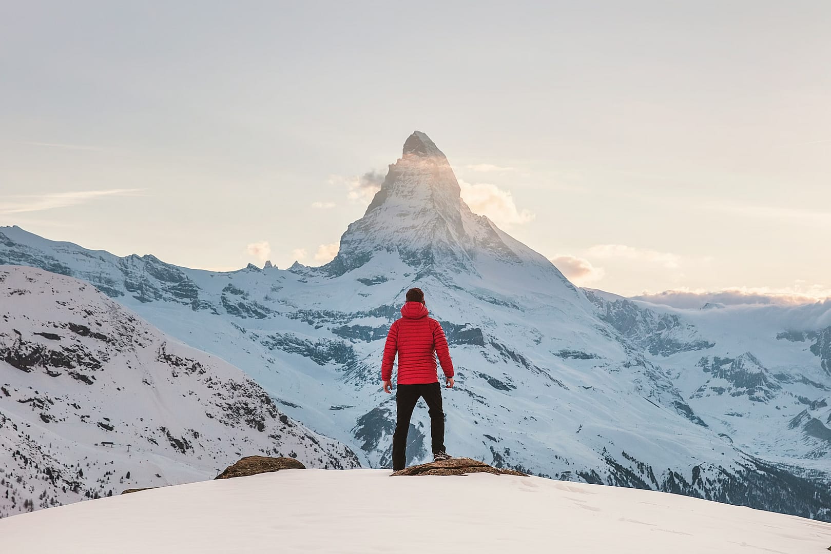 A person wearing a red jacket stands on a snowy hilltop, gazing at the iconic Matterhorn peak during sunrise or sunset. The surrounding landscape is covered in snow, with the mountain illuminated by soft light.