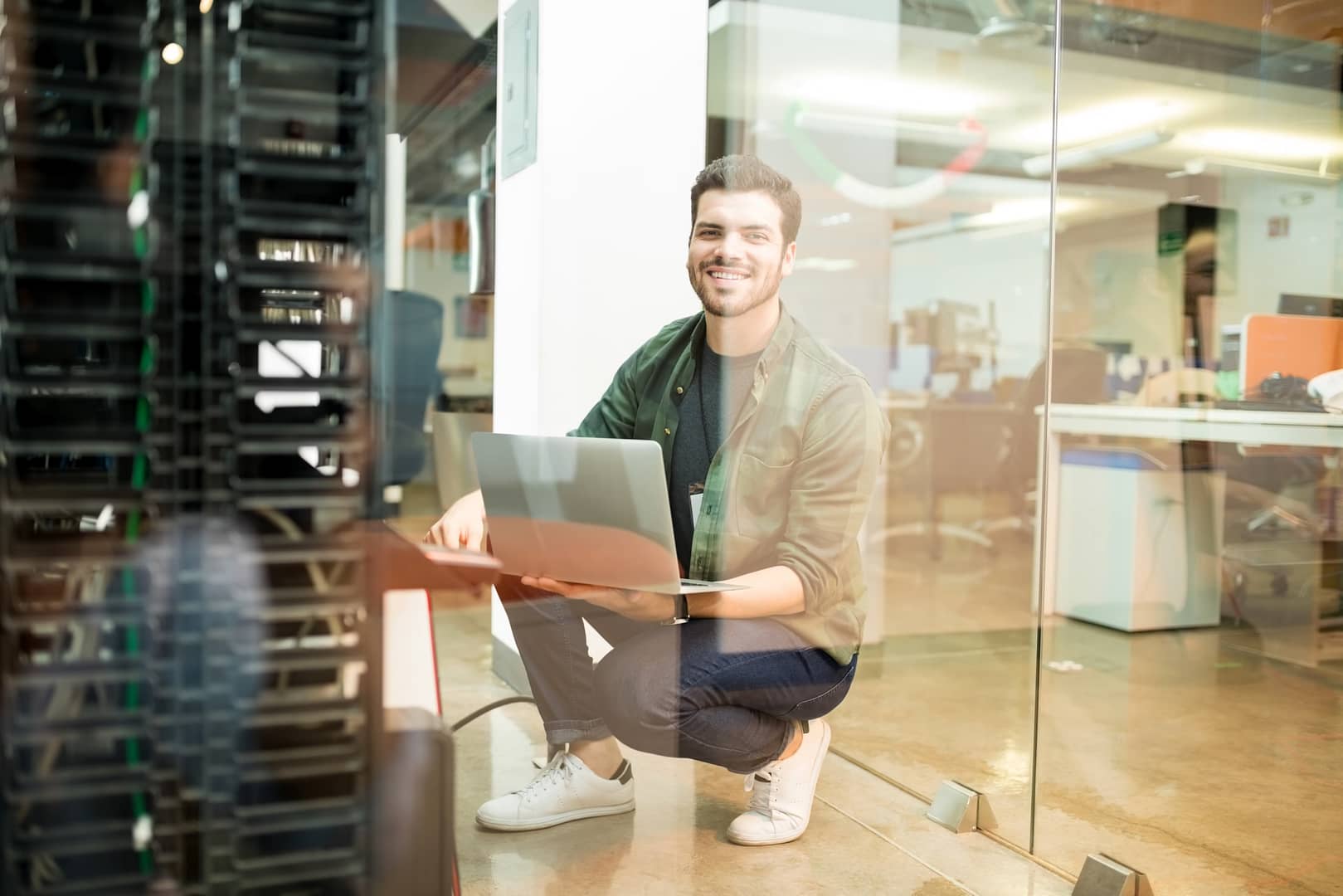 A cheerful young male network engineer with a laptop, working in a modern data centre surrounded by server racks.