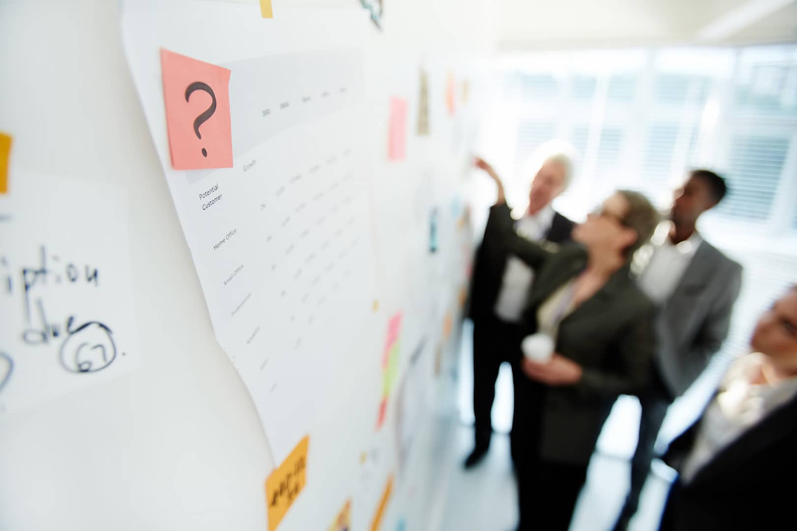 A group of professionals reviewing charts and sticky notes on a whiteboard, discussing project details in a bright office environment.